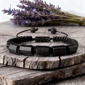 May include: Black beaded bracelet featuring rectangular black stones, set against a rustic wooden surface. The bracelet is accented with black cord and small, dark beads. A bundle of lavender is in the background.