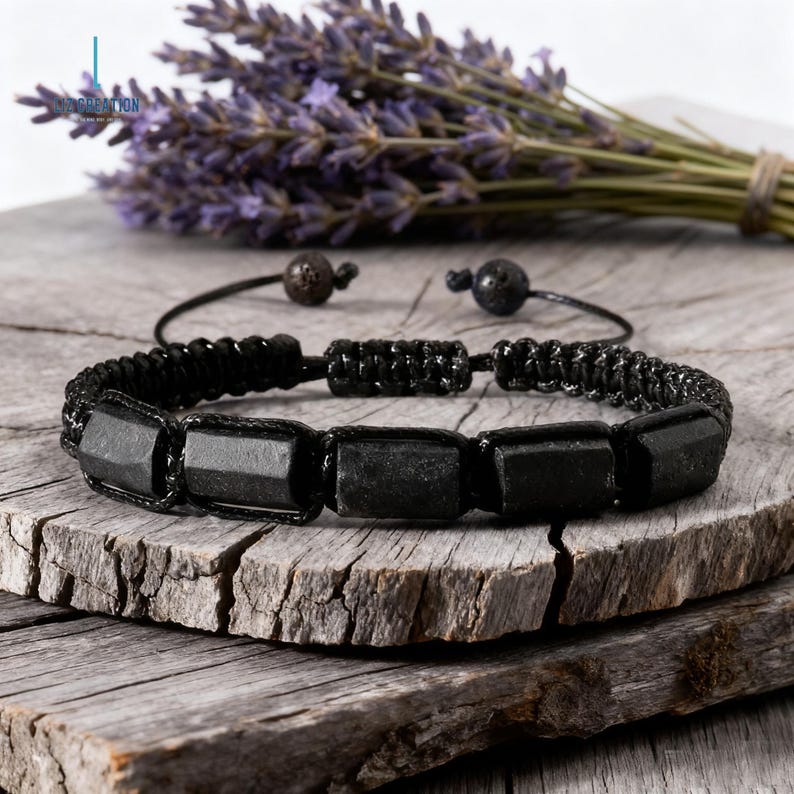 May include: Black beaded bracelet featuring rectangular black stones, set against a rustic wooden surface. The bracelet is accented with black cord and small, dark beads. A bundle of lavender is in the background.