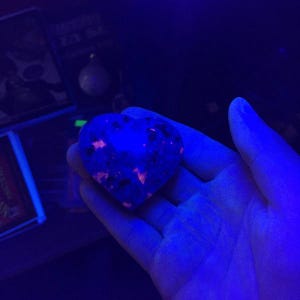 Puede incluir: Una piedra en forma de coraz&oacute;n brilla con un tono azul vibrante, salpicada de inclusiones naranjas y negras. La piedra se sostiene en una mano, iluminada por una luz azul. El fondo es oscuro, con indicios de otros objetos.