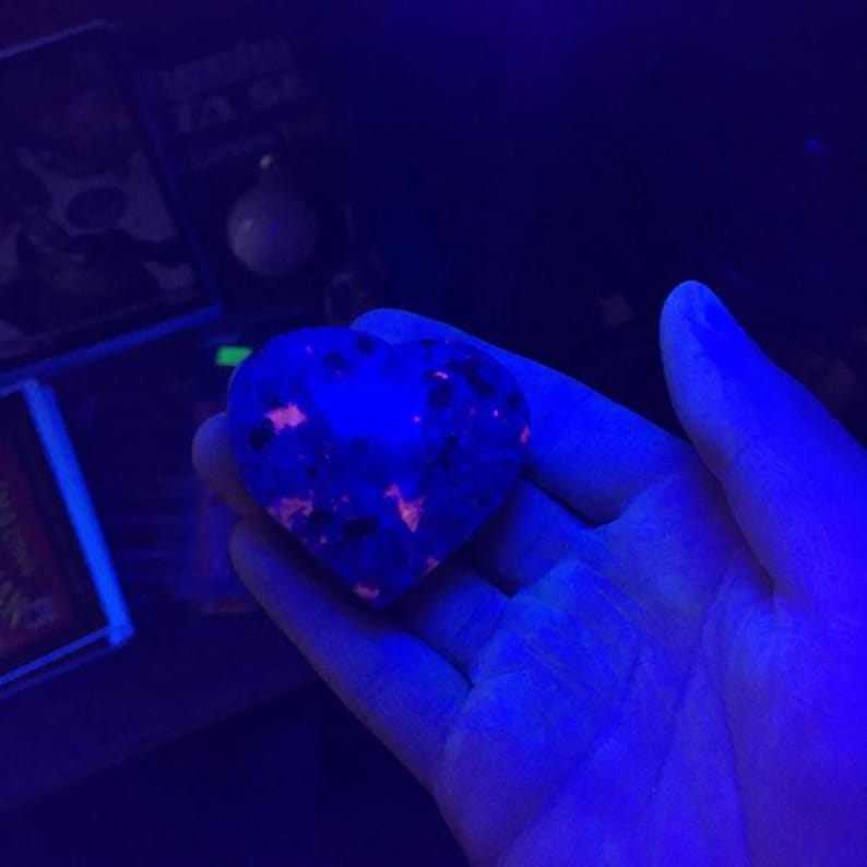 Puede incluir: Una piedra en forma de coraz&oacute;n brilla con un tono azul vibrante, salpicada de inclusiones naranjas y negras. La piedra se sostiene en una mano, iluminada por una luz azul. El fondo es oscuro, con indicios de otros objetos.