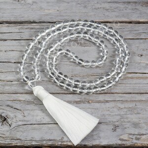 Op de afbeelding: Ketting van heldere kwarts kristallen kralen mala met een witte kwast. De kralen zijn aan elkaar geregen en vormen een cirkelvormig patroon. De ketting ligt op een verweerd, grijs houten oppervlak. De tekst "LIZ-CREATION" staat in de linkerbovenhoek.