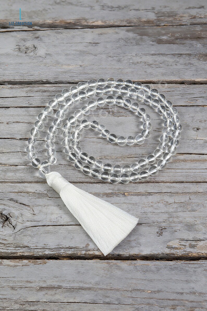 Op de afbeelding: Ketting van heldere kwarts kristallen kralen mala met een witte kwast. De kralen zijn aan elkaar geregen en vormen een cirkelvormig patroon. De ketting ligt op een verweerd, grijs houten oppervlak. De tekst "LIZ-CREATION" staat in de linkerbovenhoek.