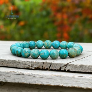 Pu&ograve; includere: Un braccialetto di perline turchesi dall'aspetto di pietra naturale. Le perline rotonde hanno un motivo screziato blu e oro. Il braccialetto &egrave; esposto su una superficie di legno invecchiato. Il testo "LIZ CREATION" &egrave; visibile in alto a sinistra.