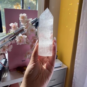 Puede incluir: Una punta de cristal clara y blanca con una superficie lisa y pulida. El cristal tiene forma de pir&aacute;mide con una punta puntiaguda y una base plana. Se sostiene en una mano, con el fondo desenfocado.