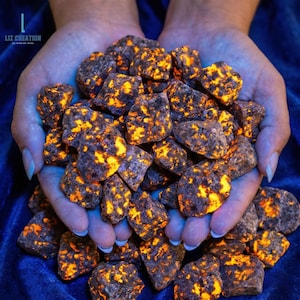 May include: A handful of rough, dark gray rocks with vibrant orange and yellow inclusions. The rocks are held in cupped hands against a dark blue background. The text "LIZ CREATION" is visible in the upper left corner.