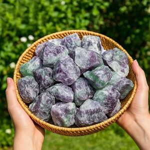 Puede incluir: Una cesta redonda de mimbre llena de cristales de fluorita en bruto. Los cristales muestran una mezcla de tonos morados, verdes y claros, con una textura natural y sin pulir. La cesta est&aacute; sostenida por dos manos sobre un fondo verde borroso.