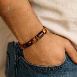 May include: A brown and gold tiger eye beaded bracelet. The bracelet is worn on a wrist, with the beads appearing to be rectangular in shape. The bracelet is set against a background of a person wearing a white shirt and blue jeans.