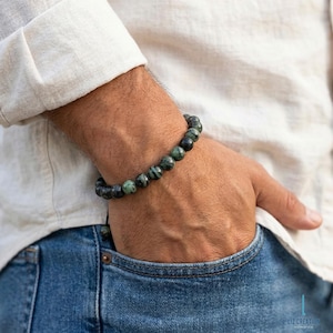 May include: A dark green beaded bracelet with black accents. The bracelet is made of round beads and is worn on the wrist. The person is wearing a white shirt and blue jeans.