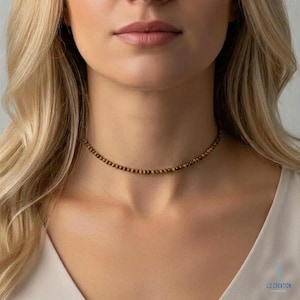 Puede incluir: Un collar gargantilla con pequeñas cuentas redondas marrones y doradas. El collar se lleva alrededor del cuello. La imagen muestra el collar sobre un fondo neutro.
