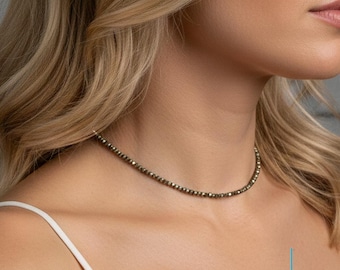 May include: A fine choker necklace made of small, round, metallic beads. The necklace sits around the neck, displaying its gentle shimmer. The background is a soft, neutral tone, accentuating the jewellery. The brand name "LIZ CREATION" is visible.