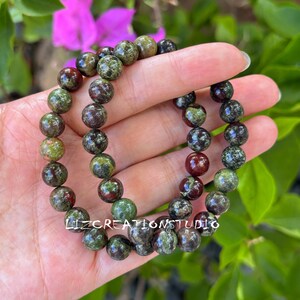 Puede incluir: Tres pulseras de cuentas con piedras verdes, marrones y rojas. Las pulseras est&aacute;n hechas de cuentas redondas y pulidas. Las pulseras se sostienen en una mano, con un fondo de follaje verde y una flor rosa. El texto "LIZCREATIONSTUDIO" es visible.