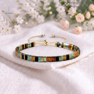 Pode incluir: Uma pulseira com um padrão repetitivo de contas retangulares em preto, bronze metálico e azul-petróleo iridescente. A peça inclui um cordão ajustável claro com um fecho de conta deslizante e ponteiras de metal prateado. O texto diz LIZ CREATION.