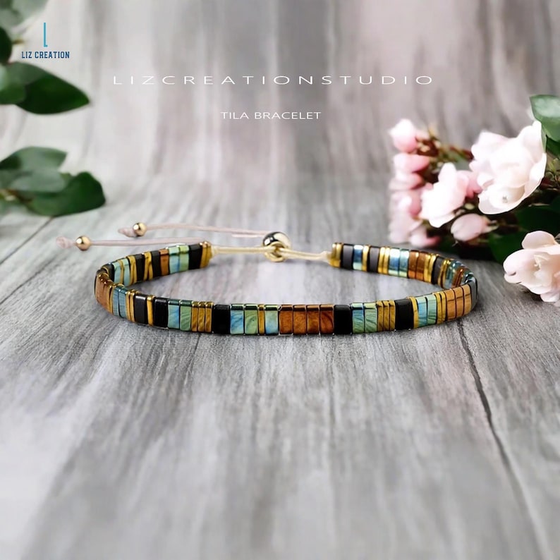 Op de afbeelding: Een veelkleurige kralen armband met een gouden sluiting en een lichtroze koord. De armband heeft een patroon van blauwe, groene, zwarte en bruine kralen. De tekst "TILA BRACELET" is zichtbaar in de afbeelding.