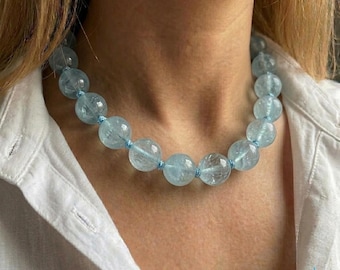 May include: A necklace featuring large, round, light blue aquamarine beads strung on a light blue cord. The beads are translucent and have a polished finish. The necklace is worn against a white shirt.