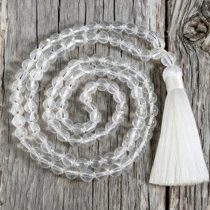 Op de afbeelding: Heldere kwarts kristallen mala kralen met een witte kwast. De kralen zijn rond en aan elkaar geregen, waardoor een spiraal ontstaat. De achtergrond is een verweerd, grijs houten oppervlak. De tekst "LIZ CREATION" staat in de linkerbovenhoek.