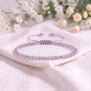 May include: A light purple beaded bracelet featuring small, round, translucent amethyst-colored beads and two metallic silver-toned spacer beads. The bracelet has an adjustable cord closure with a sliding knot mechanism and two small beads at the ends.