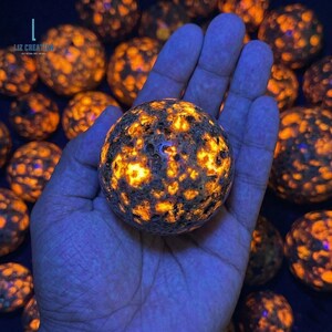 May include: A hand holds a glowing, spherical stone with a dark, textured surface and bright orange and yellow inclusions. Other similar spheres are in the background. The image has a blue tint.