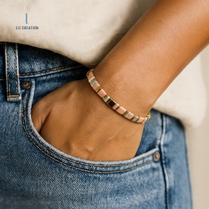 Könnte beinhalten: Ein Armband mit Perlen aus rosa, goldfarbenen, hellblauen und braunen rechteckigen Perlen. Das Armband wird am Handgelenk getragen, die Hand steckt in einer blauen Jeans-Tasche. Das Armband hat einen goldfarbenen Verschluss.
