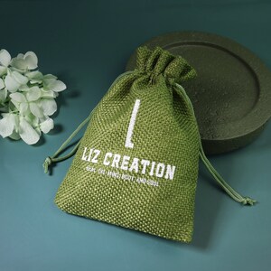 Op de afbeelding: Een groene jute trekkoordtas met witte tekst die "Liz Creation Heal the Mind, Body and Soul" luidt.