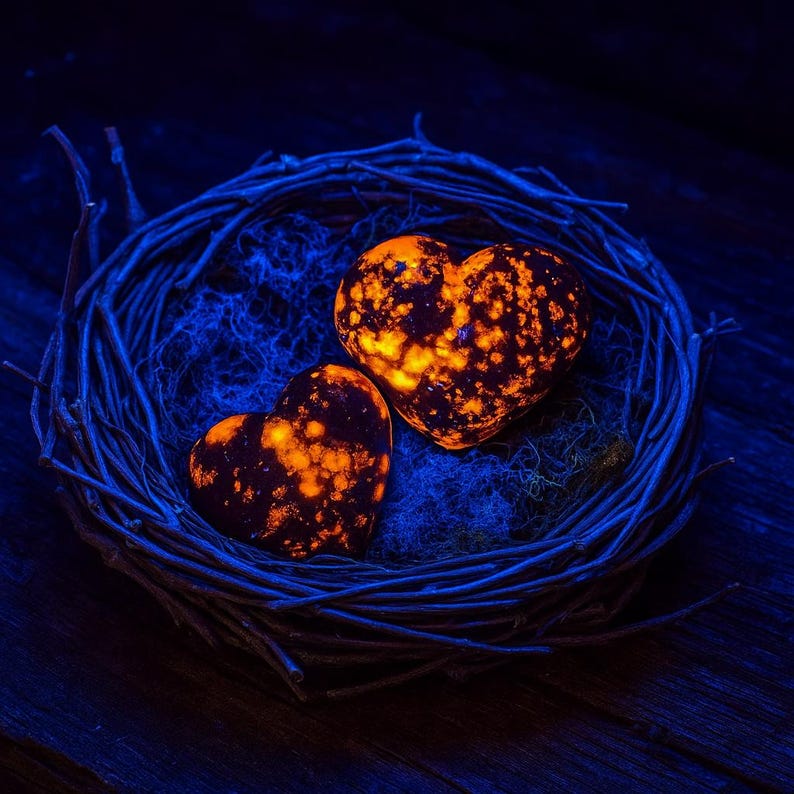 Puede incluir: Dos piedras en forma de coraz&oacute;n con patrones naranjas y negros descansan en un nido tejido. El nido est&aacute; hecho de ramitas azul oscuro y descansa sobre una superficie azul oscuro. Las piedras brillan, creando un contraste llamativo.