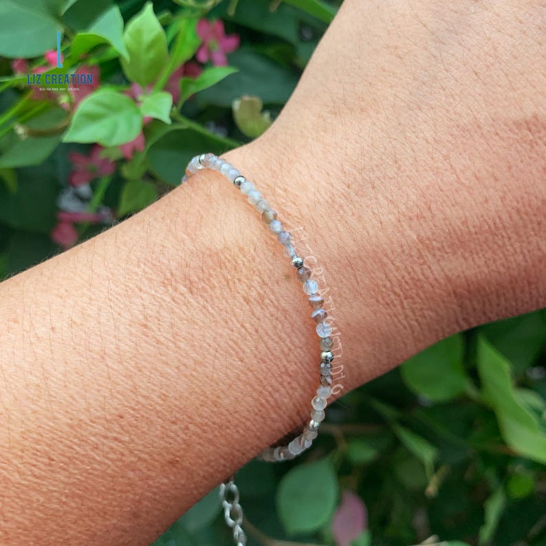 May include: A delicate bracelet made with small, light gray and silver beads. The bracelet is on a person's wrist.