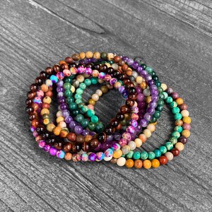Op de afbeelding: Een stapel kleurrijke kralenarmbanden, met verschillende edelsteenkleuren zoals paars, groen, bruin en oranje. De armbanden zijn gemaakt van kleine, ronde kralen en zijn in een cirkelvormige opstelling gerangschikt.