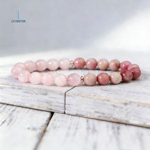 Puede incluir: Un brazalete de cuentas con piedras preciosas rosas y rosadas. El brazalete tiene peque&ntilde;os detalles met&aacute;licos y se muestra sobre una superficie de madera blanca y desgastada. El texto "LIZ CREATION" es visible en la esquina superior izquierda.