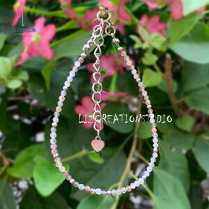 May include: A delicate silver bracelet with a heart charm, featuring small, iridescent beads in shades of white, pink, and gray.