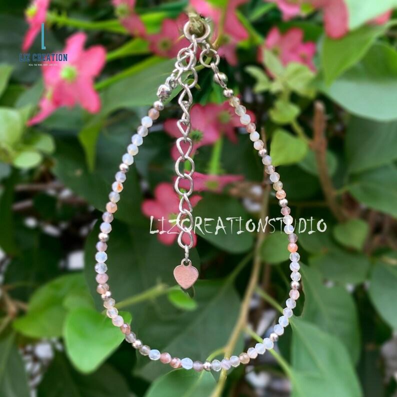 May include: A delicate silver bracelet with a heart charm, featuring small, iridescent beads in shades of white, pink, and gray.