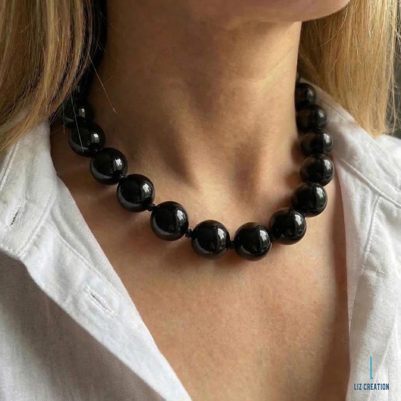 May include: A black beaded necklace with large, round beads. The necklace is worn against a white collared shirt. The beads have a glossy finish and are strung together. The necklace is a statement piece.