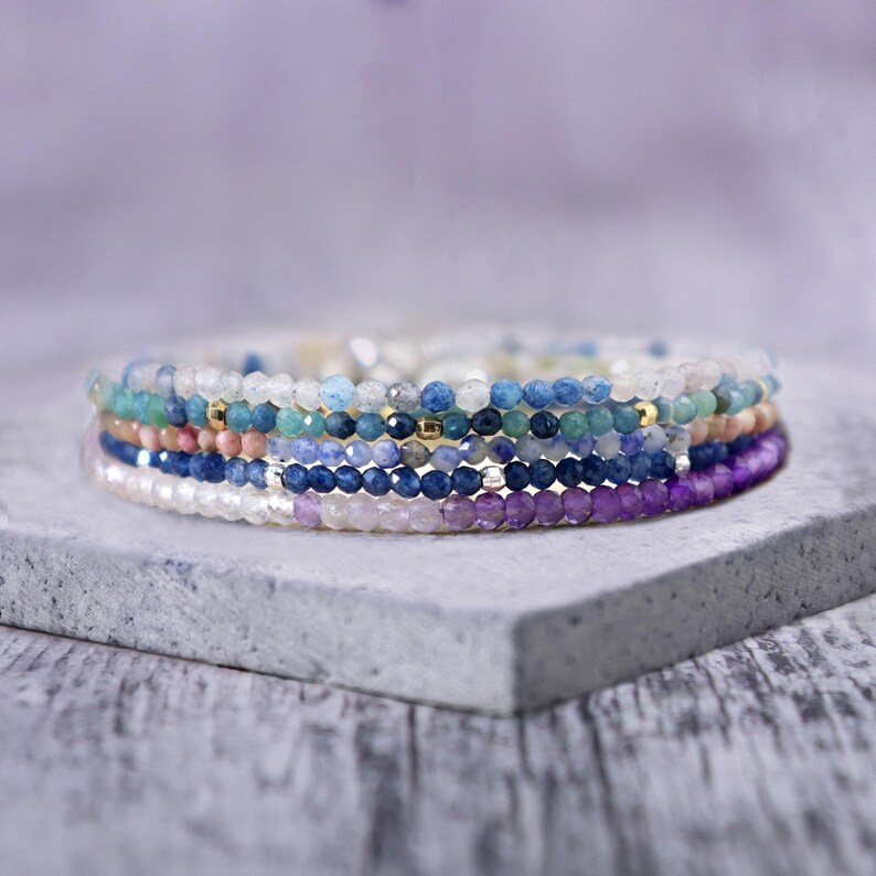 May include: A stack of colorful gemstone bracelets in shades of blue, purple, and pink. The bracelets are made of small, round beads and are layered on top of each other.