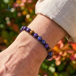 Può includere: Un braccialetto di perline con una miscela di pietre di occhio di tigre viola e marroni. Il braccialetto è indossato su un polso, con le pietre di circa 0,8 cm di diametro. È un gioiello.