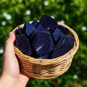 May include: A woven basket filled with dark blue gemstones. The gemstones have a sparkling, glittery appearance. The basket is made of light brown woven material. The background is a blurred green.