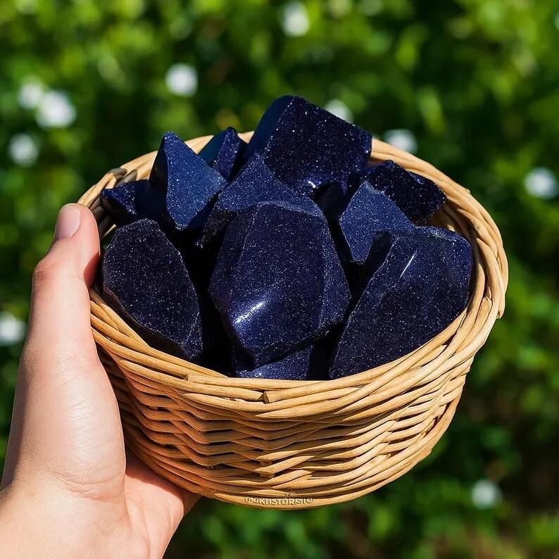 May include: A woven basket filled with dark blue gemstones. The gemstones have a sparkling, glittery appearance. The basket is made of light brown woven material. The background is a blurred green.