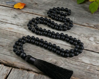 May include: Black beaded mala necklace with a long black tassel. The necklace is made of small, round beads and is displayed on a weathered wooden surface. The image also includes a matching bracelet.