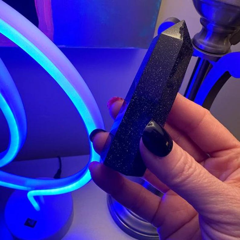 May include: A dark blue, glittery crystal point is held in a hand. The crystal has a faceted, pointed top and a rectangular shape. The background is a blurred display of blue and purple neon lights.