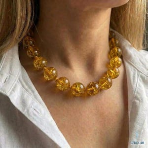 Puede incluir: Un collar que presenta una sola hilera de cuentas redondas y translúcidas de color amarillo dorado con una textura interna agrietada. Las cuentas son de tamaño uniforme y están ensartadas formando un círculo alrededor del cuello. El texto LIZ CREATION es visible.