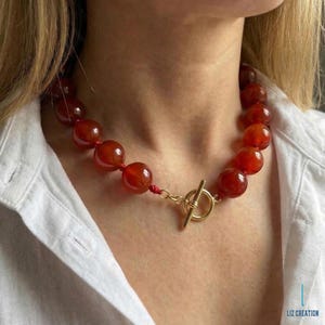 May include: A red beaded necklace with a gold-colored clasp. The necklace features large, round, polished beads. The clasp is a T-bar style. The necklace is worn against a white shirt.