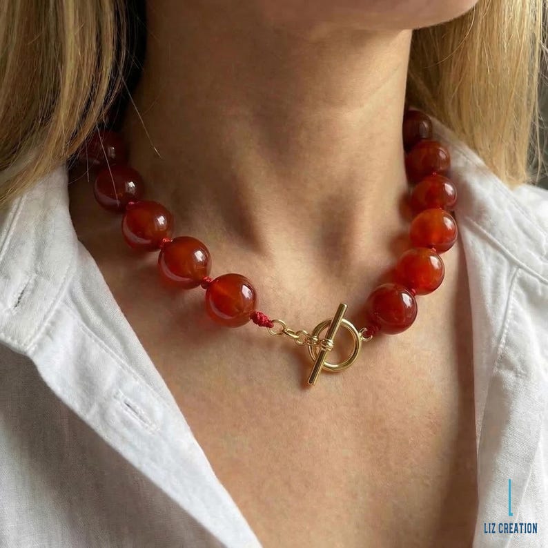 May include: A red beaded necklace with a gold-colored clasp. The necklace features large, round, polished beads. The clasp is a T-bar style. The necklace is worn against a white shirt.