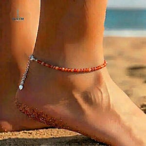 May include: Anklet featuring small, round, orange and white beads strung together. The anklet has a silver-colored chain with a heart-shaped charm. The anklet is worn on an ankle, with a beach background.