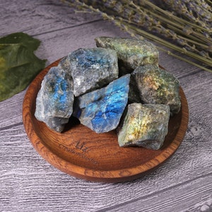 May include: A collection of raw labradorite stones with iridescent blue and green flashes, displayed in a small, round wooden bowl. The stones vary in size and shape, showcasing natural textures and colors. The bowl has the text "Liz Creation Studio" engraved on it.