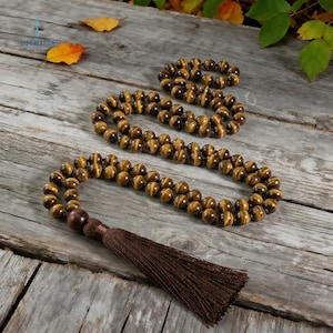 Op de afbeelding: Een set tijgeroog edelsteen gebedskralen met een bruine kwast. De kralen hebben een warme bruine kleur met gouden strepen. De kralen zijn gerangschikt op een houten oppervlak met herfstbladeren.