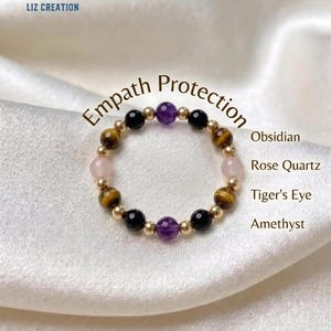 May include: A beaded ring featuring alternating round beads of purple amethyst, black obsidian, brown tiger's eye, and pale pink rose quartz, interspersed with small gold-toned metallic beads. The text "Empath Protection" is visible above the ring.