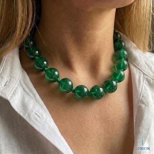 May include: A necklace featuring a single strand of large, round, translucent emerald green beads. The beads are strung together with a matching green cord, creating a uniform circular design that rests around the neck. The text LIZ CREATION is visible.