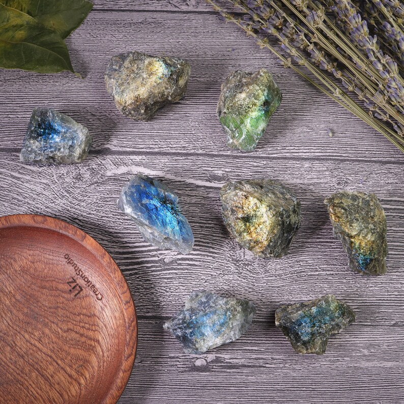 May include: An overhead shot displays several raw labradorite stones with iridescent blue, green, and gold flashes. The stones are arranged on a wooden surface, alongside a wooden bowl and sprigs of dried lavender. The stones vary in size and shape.