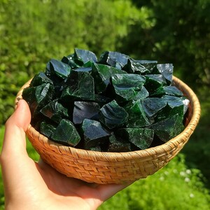 Puede incluir: Una cesta de mimbre llena de trozos de vidrio verde oscuro de forma irregular. Las piezas de vidrio tienen una superficie brillante y reflectante. La cesta se sostiene contra un fondo verde borroso, lo que sugiere un entorno al aire libre.