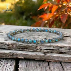 Könnte beinhalten: Ein Armband aus Labradorit-Perlen, bestehend aus kleinen, runden Perlen mit irisierenden blauen und grünen Reflexen auf grauem Grund. Das Armband wird auf einer verwitterten Holzoberfläche präsentiert. Der Text "LIZ CREATION" ist oben links zu sehen.