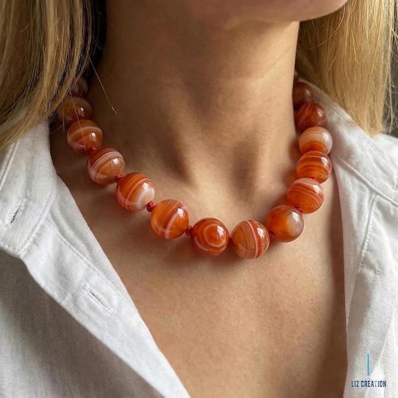 May include: A necklace with large, spherical beads in orange and white hues. The beads are strung on a red cord, creating a striking accessory. The necklace is worn against a white shirt. The beads are approximately 2 cm in diameter.