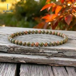 May include: A beaded bracelet with small, round beads in shades of green, brown, and reddish-orange. The bracelet is resting on a weathered wooden surface. The text "LIZ CREATION" is in the upper left corner.
