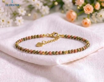 Könnte beinhalten: Ein zartes Armband mit kleinen, facettierten, runden Perlen in abwechselnden Farbtönen von Olivgrün und Altrosa. Das Schmuckstück enthält goldfarbene Metallzwischenstücke, einen Karabinerverschluss, eine verstellbare Kette und einen kleinen herzförmigen Anhänger.